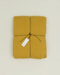The Simple Linen Quilt in Mustard by Hawkins New York is shown folded and tied with black string, featuring a white tag, and displayed on a plain light background.
