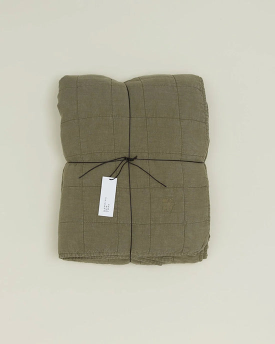 The Hawkins New York Simple Linen Quilt in Olive, featuring a grid pattern, is folded and tied with black string and a white tag, displayed on a plain light background.