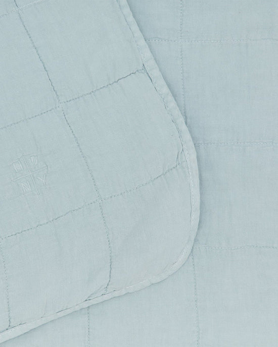 The Simple Linen Quilt in Sky by Hawkins New York features light blue stonewashed linen, a grid stitch pattern, rounded edges, and a small embroidered logo on the corner—offering timeless style for any room.