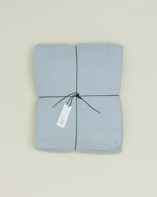 The Hawkins New York Simple Linen Quilt in Sky, neatly folded and tied with black string, features a white tag and rests on a plain light background.