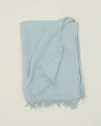 The Simple Linen Throw in Sky by Hawkins New York is shown neatly folded with fringed edges on a plain, light background.