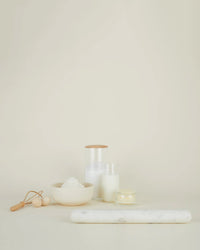 A minimalistic scene with a Hawkins New York Simple Marble Rolling Pin, a bowl of flour, a small glass jar of milk, a lit candle, a storage jar, and a wooden-handled dough cutter on a light beige background.