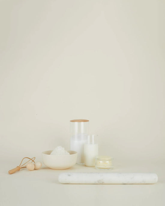 A minimalistic scene with a Hawkins New York Simple Marble Rolling Pin, a bowl of flour, a small glass jar of milk, a lit candle, a storage jar, and a wooden-handled dough cutter on a light beige background.