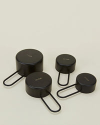 Four Simple Measuring Cups in Black by Hawkins New York—labeled 1 cup, 1/2 cup, 1/3 cup, and 1/4 cup—are arranged on a light surface.
