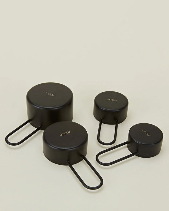 Four Simple Measuring Cups in Black by Hawkins New York—labeled 1 cup, 1/2 cup, 1/3 cup, and 1/4 cup—are arranged on a light surface.