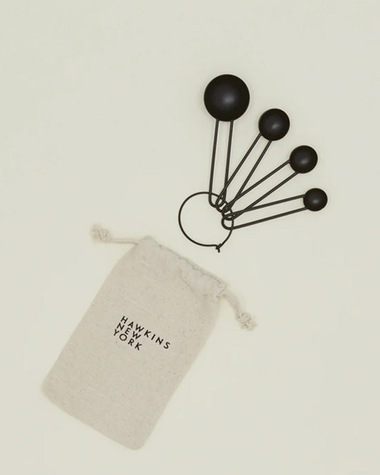 Four Simple Measuring Spoons in Black by Hawkins New York are held on a ring and shown beside a beige drawstring pouch labeled with the brand’s name.