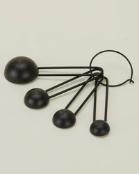 The Simple Measuring Spoons in Black by Hawkins New York are a set of four stainless steel spoons in different sizes, connected by a metal ring and shown on a light beige background.