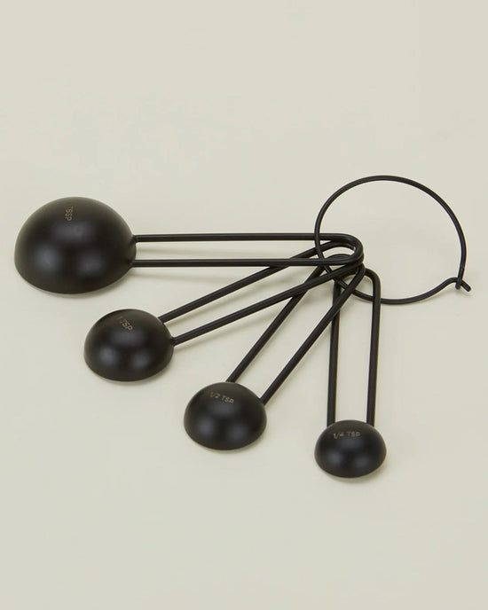 The Simple Measuring Spoons in Black by Hawkins New York are a set of four stainless steel spoons in different sizes, connected by a metal ring and shown on a light beige background.