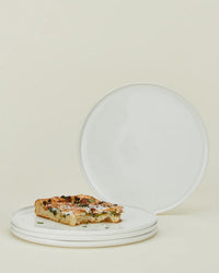 A stack of three Simple Porcelain Dinner Plates from Hawkins New York holds a rectangular slice of pizza, while a fourth plate from the set of 4 is propped upright behind them against a light beige background.