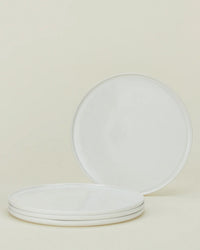 Four Simple Porcelain Dinner Plates by Hawkins New York are displayed on a light beige background—three stacked horizontally and one propped vertically behind—highlighting their elegant fine porcelain style.
