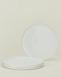 Four Simple Porcelain Dinnerware plates from Hawkins New York are displayed on a neutral background; three are stacked and one stands upright behind, highlighting this minimalist 16-piece set.