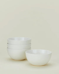 Four white porcelain bowls from the Hawkins New York Simple Porcelain Dinnerware - 16 Piece Set are displayed: three stacked on the left, one in front on the right, all against a light background.