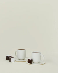 The Simple Porcelain Espresso Cups & Saucers - Set of 2 by Hawkins New York are shown on a plain light background, each on its saucer and paired with a chocolate brownie and a sugar-dusted brownie.