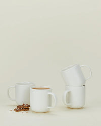 Four Hawkins New York Simple Porcelain Mugs—set of 4—are shown on a light background; two are stacked, one holds coffee, and one is empty with chocolate pieces scattered nearby.