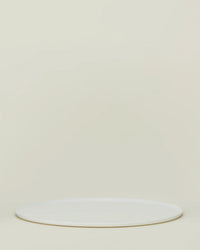 The Simple Porcelain Serving Platter by Hawkins New York, a round white porcelain piece, rests on a light beige background with empty space above, reflecting the brand’s minimalist elegance.