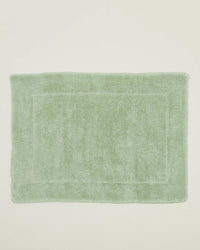 The Simple Terry Bath Mat in Sage by Hawkins New York is a rectangular, soft cotton bath mat with a subtle border design, shown on an off-white background.