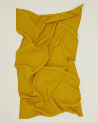 The Simple Waffle Bath Sheet in Mustard by Hawkins New York is spread on a flat surface, loosely arranged with soft folds and textured wrinkles, showcasing its signature waffle weave.
