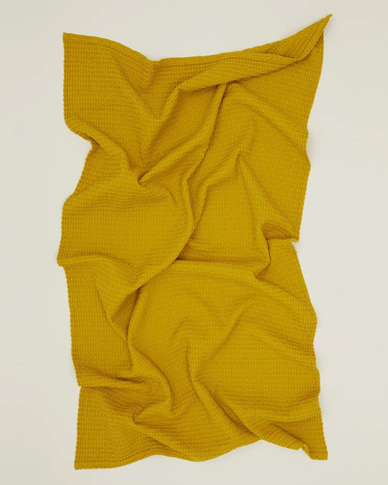 The Simple Waffle Bath Sheet in Mustard by Hawkins New York is spread on a flat surface, loosely arranged with soft folds and textured wrinkles, showcasing its signature waffle weave.