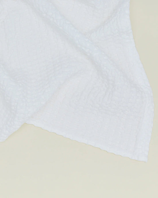 A close-up of the Hawkins New York Simple Waffle Bath Sheet in White, showing its textured grid pattern reminiscent of waffle towels, draped against a light beige background.