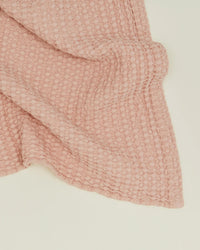 The Hawkins New York Simple Waffle Hand Towel in Blush features a textured, waffle-like pattern and light pink hue, crafted in Portugal to add elegance and charm to any space. Draped on a plain, light background.