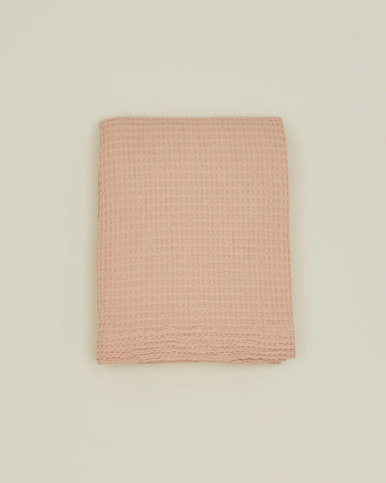 A folded, blush waffle-textured towel rests on a plain off-white background, evoking the delicate weave of the Hawkins New York Simple Waffle Shower Curtain.