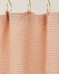 The Simple Waffle Shower Curtain in Blush by Hawkins New York, made from textured cotton, is shown hanging on three gold hooks against a neutral background.