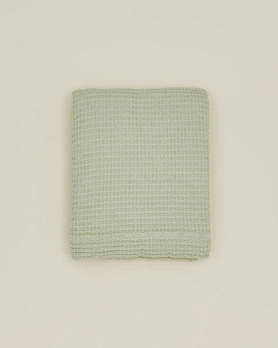 A folded Simple Waffle Shower Curtain in Sage by Hawkins New York, featuring a waffle texture, is centered on a plain beige background.