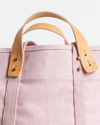 Close-up of the IMMODEST COTTON Large East West Tote in Bubblegum, an oversized light pink bag with tan leather handles, brass studs, and reinforced stitching shown against a white background.