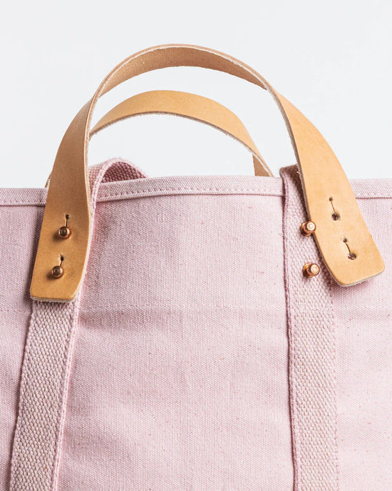 Close-up of the IMMODEST COTTON Large East West Tote in Bubblegum, an oversized light pink bag with tan leather handles, brass studs, and reinforced stitching shown against a white background.
