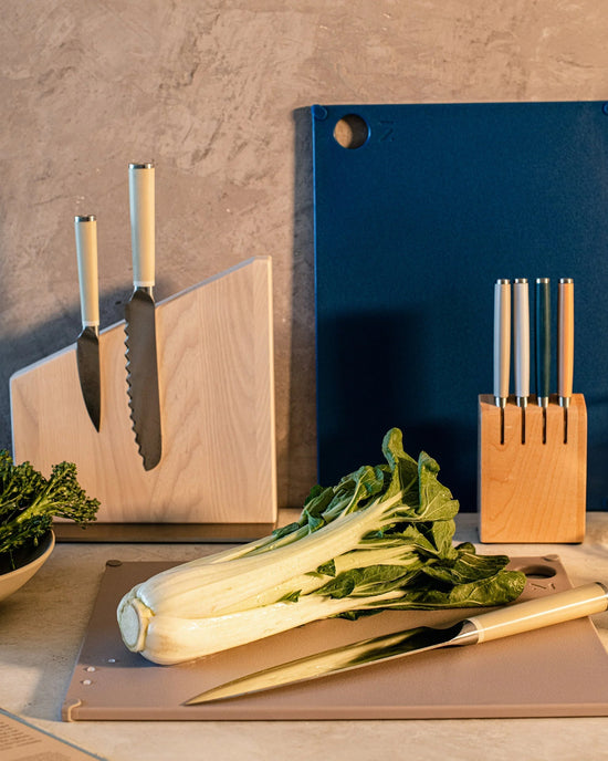 A kitchen counter with bok choy on a BPA-free cutting board, a knife beside it, two knife blocks, a bowl of greens from recycled scraps, and the Material (grippy) reBoard® | Lapis leaning against the wall.
