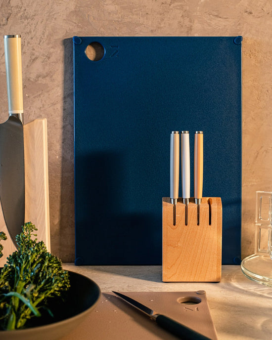 The Material (grippy) reBoard® | Lapis, a BPA-free cutting board, stands upright behind a wooden knife block with three knives; a chefs knife, glass of water, and vegetables sit on the countertop.