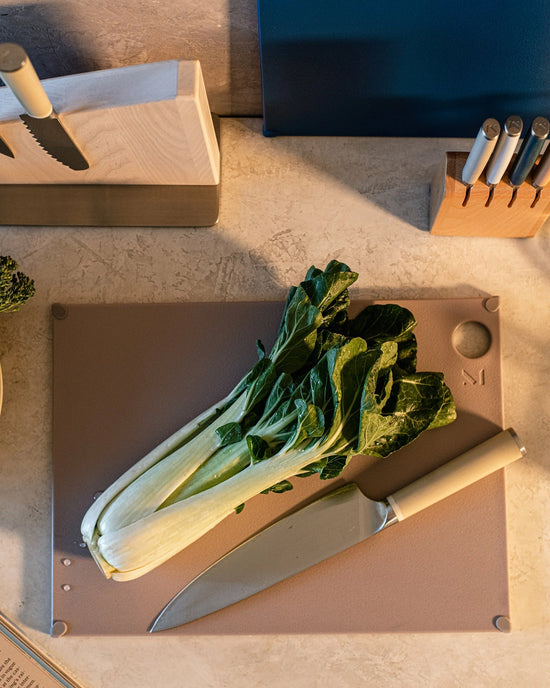 A bunch of bok choy and a kitchen knife rest on The (grippy) reBoard® | Taffy cutting board by Material, with knife blocks and utensils in the background.