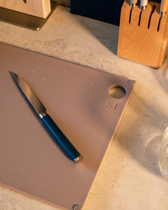 The (grippy) reBoard® | Taffy by Material sits on a countertop with water droplets, holding a kitchen knife beside a wooden knife block.