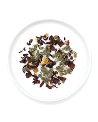 On a white plate sits an inviting blend of dried herbs, flower petals, and fruit, evoking the caffeine-free Raspberry Hibiscus Tea by Onyx Coffee Lab—an antioxidant-rich tea available in 15 sachets.