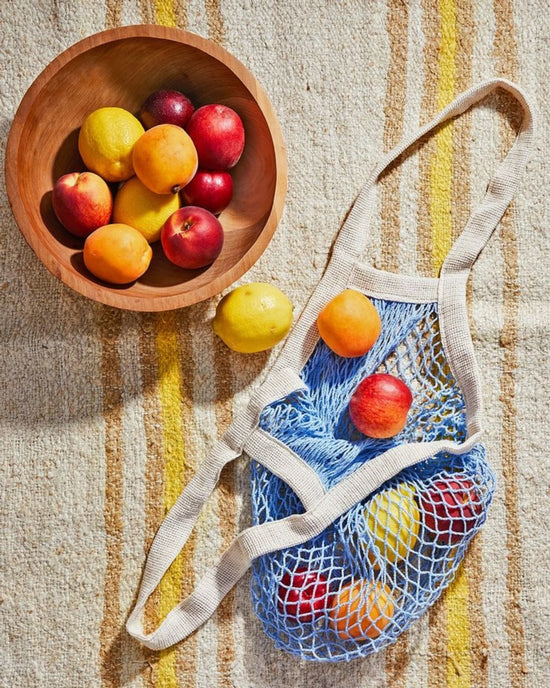 A wooden bowl of assorted fruit sits beside PILLOWPIA’s The French Market Bag No.2 in French Blue, filled with similar fruit, on a striped fabric surface.