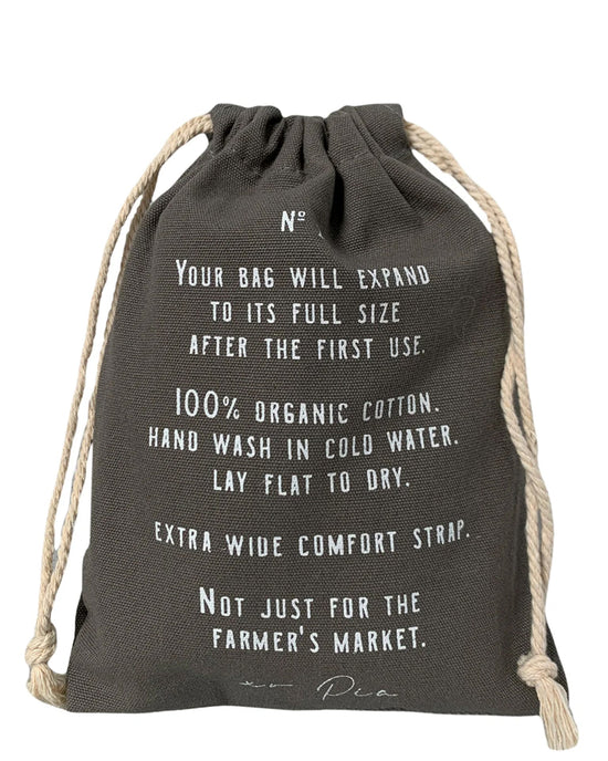 The French Market Bag No.2 in Iron by PILLOWPIA is a black drawstring bag with white care instructions, made from 100% organic cotton, featuring an extra wide comfort strap and recommended cold hand washing.