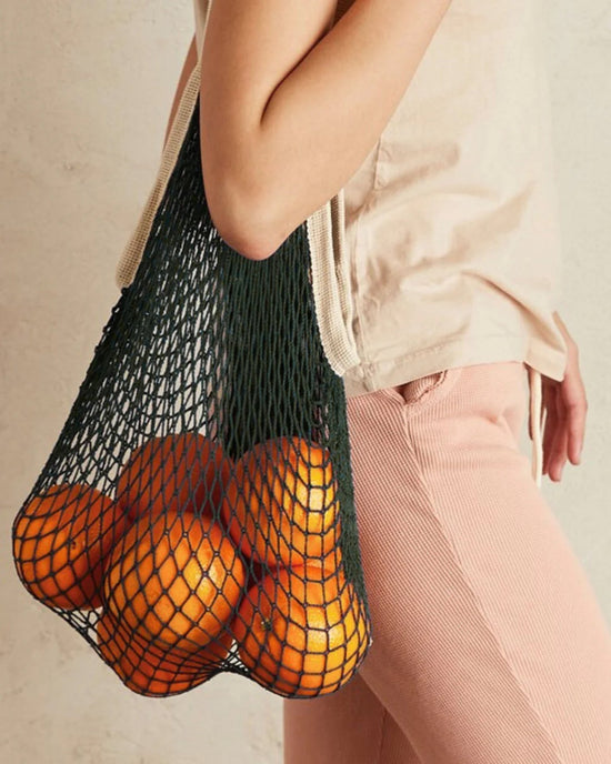 A person carries PILLOWPIAs The French Market Bag No.2 In Iron, filled with oranges, and wears a beige shirt with light pink pants.