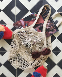 The French Market Bag No.2 In Natural by PILLOWPIA sits on a black-and-white tile floor with round sunglasses, a fashion magazine, and red espadrilles featuring blue-and-white tassels.