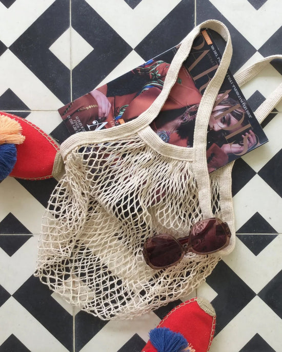 The French Market Bag No.2 In Natural by PILLOWPIA sits on a black-and-white tile floor with round sunglasses, a fashion magazine, and red espadrilles featuring blue-and-white tassels.