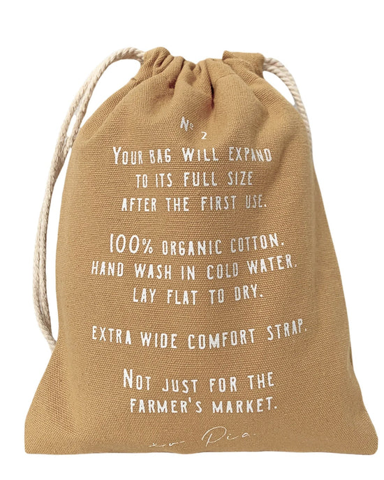 The French Market Bag No.2 in Wheat by PILLOWPIA is a tan organic cotton bag with white print, wide strap, and drawstring closure. Hand wash only—ideal for errands and outings beyond the farmers market.
