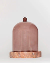 The Cloche + Stone in Nude by Poured New York features a brown glass cloche cover with a round knob resting on a circular stone coaster—a chic candle accessory for any space.