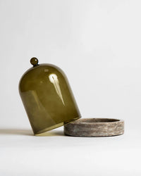 The Cloche + Stone in Olive by Poured New York features a green-tinted glass cloche tilted beside its round stone base, shown against a plain white background.