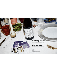 A set table with a wine bottle, plates, glasses, a candle, Polaroid photos, and the Nothing Fancy cookbook by Random House—perfect inspiration for easy entertaining recipes.