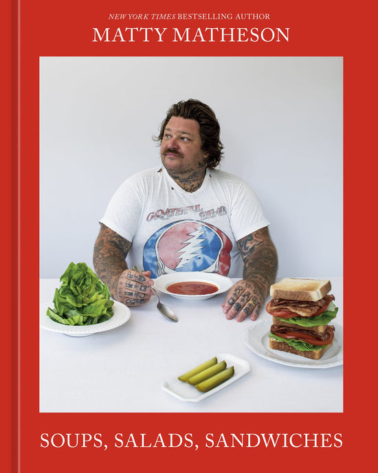 A tattooed man sits at a table enjoying tomato soup, salads, pickles, and a stacked sandwich. On the table is Soups, Salads, Sandwiches by Random House.