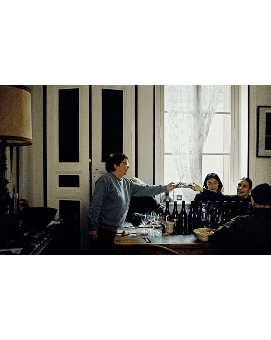 Four people sit around a table with Spilled Natural Winemakers Stories & Recipes by Random House, enjoying wine, as one person stands to hand an item to someone near a window with sheer curtains.