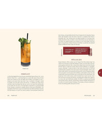 A copy of The Encyclopedia of Cocktails by Random House lies beside a Pimm’s Cup garnished with mint and cucumber—a refreshing scene for cocktail enthusiasts.