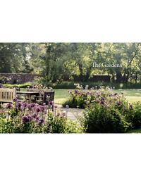 Wooden benches and tables rest on a stone patio among flowers and greenery in a sunlit English-style garden, highlighted by “The New Romantic Garden” from Random House in the background.