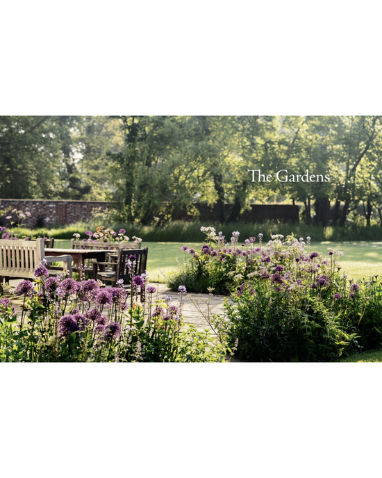 Wooden benches and tables rest on a stone patio among flowers and greenery in a sunlit English-style garden, highlighted by “The New Romantic Garden” from Random House in the background.