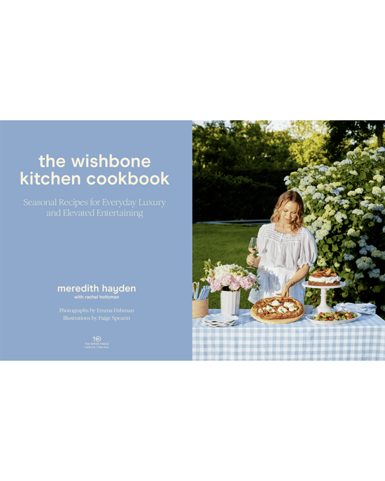 A woman in a light dress sets food on an outdoor table by flowers. On the left is The Wishbone Kitchen Cookbook, published by Random House, featuring dinner party recipes by Meredith Hayden and contributor credits.