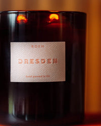 A dark glass candle jar labeled ROEN Le Grand Dresden, hand poured in CA with coconut soy wax, is lit against a soft orange background.